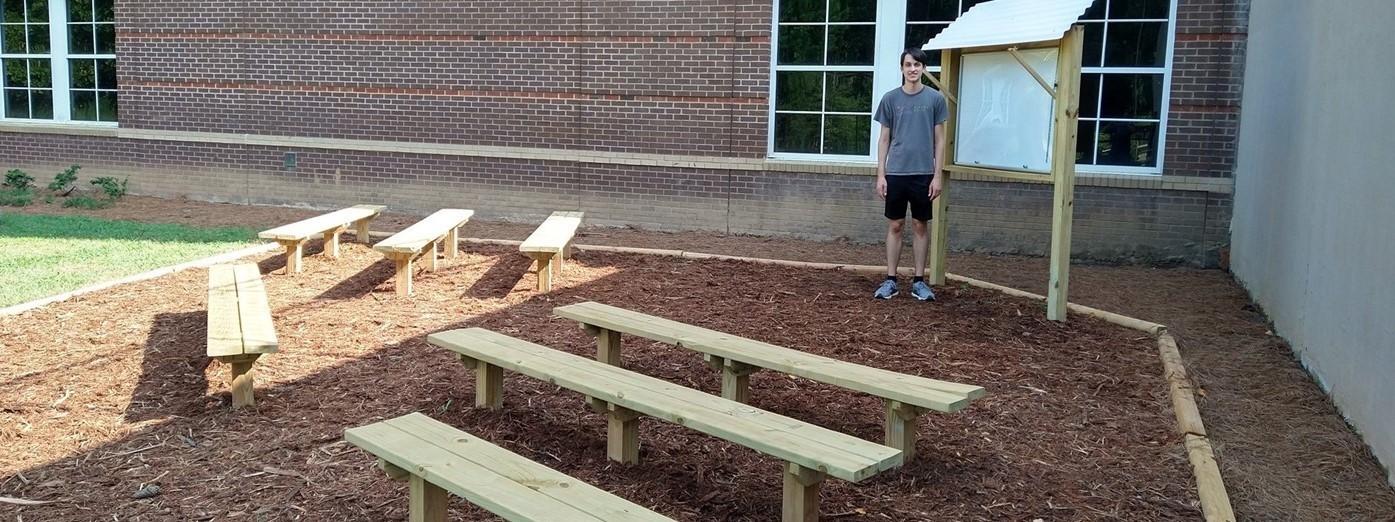 Picture of a boy standing next to an outdoor space where he built a whiteboard and 7 benches.